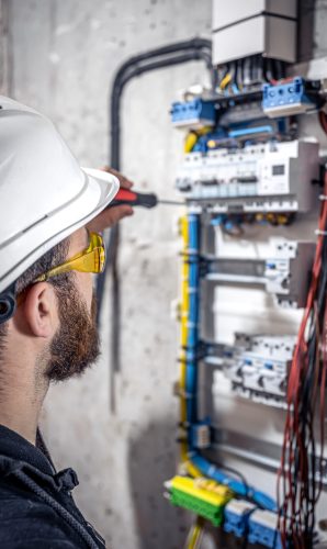 A male electrician works in a switchboard with an electrical connecting cable, connects the equipment with tools.