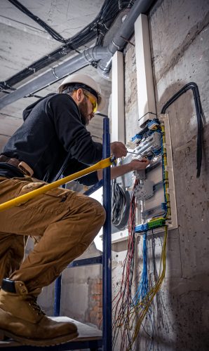 A male electrician works in a switchboard with an electrical connecting cable, connects the equipment with tools.